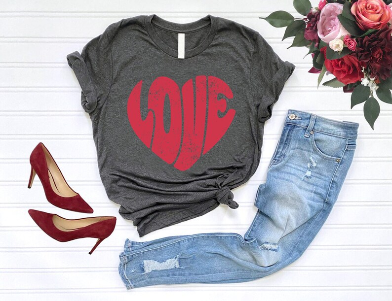 May include: A dark gray t-shirt with a red heart-shaped graphic that says "LOVE" in a distressed font. The shirt is paired with light blue jeans and red heels.
