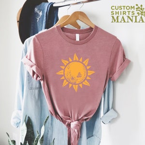 Puede incluir: Una camiseta rosa con un gr&aacute;fico de sol amarillo con mariposas y detalles florales. El texto "CUSTOM SHIRTS MANIA" est&aacute; en la esquina superior derecha de la imagen.