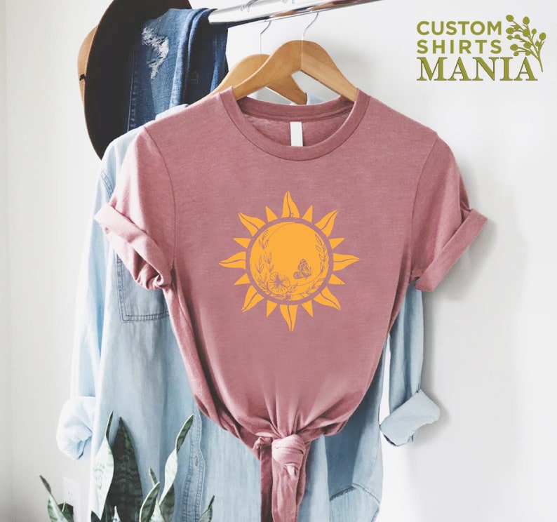 Puede incluir: Una camiseta rosa con un gr&aacute;fico de sol amarillo con mariposas y detalles florales. El texto "CUSTOM SHIRTS MANIA" est&aacute; en la esquina superior derecha de la imagen.