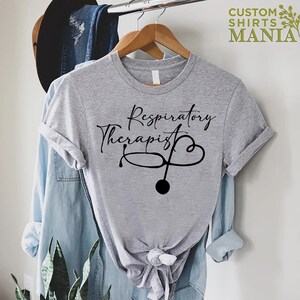 May include: Gray t-shirt with the text "Respiratory Therapist" in black cursive lettering and a stethoscope with a heart design.