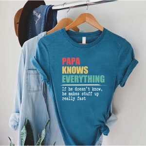 May include: Teal t-shirt with the text "PAPA KNOWS EVERYTHING" in red, yellow, and green. Below the text is the quote, "If he doesn't know, he makes stuff up really fast."