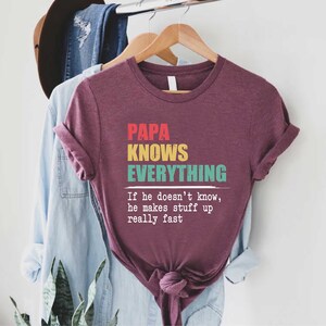May include: A maroon t-shirt with the text "PAPA KNOWS EVERYTHING" in red, yellow, and teal. The phrase "If he doesn't know, he makes stuff up really fast" is printed below the main text.