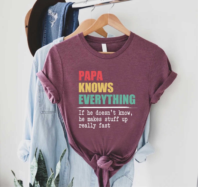 May include: A maroon t-shirt with the text "PAPA KNOWS EVERYTHING" in red, yellow, and teal. The phrase "If he doesn't know, he makes stuff up really fast" is printed below the main text.