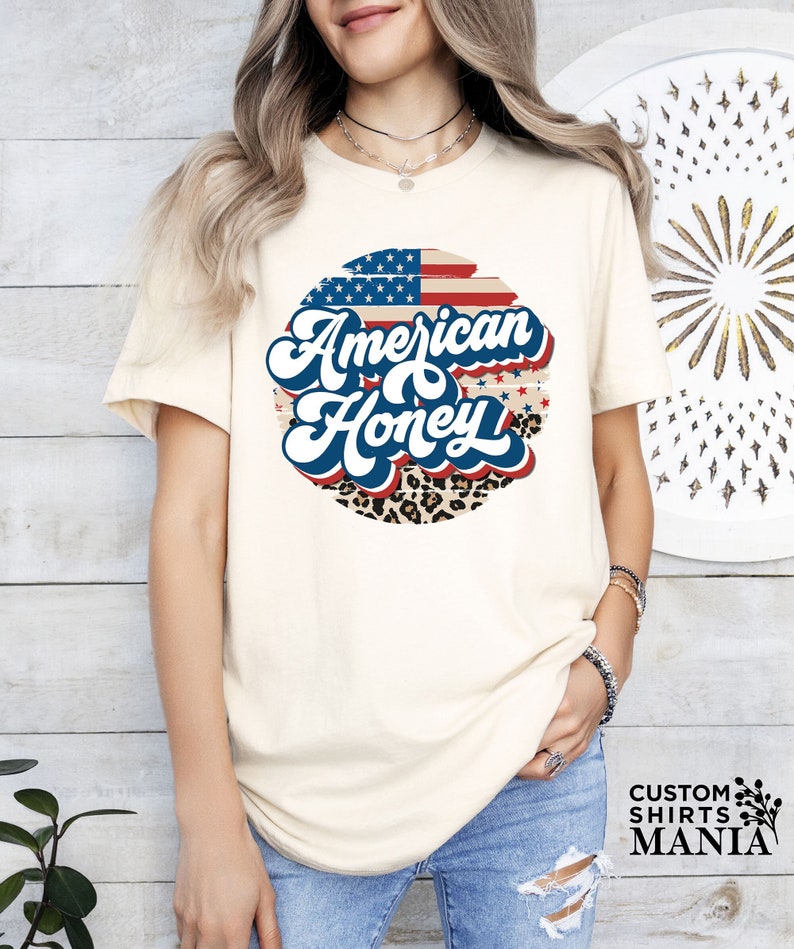 May include: A white t-shirt with a circular graphic featuring an American flag, leopard print, and the text "American Honey" in a retro font.
