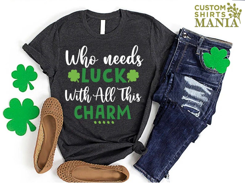May include: A dark gray t-shirt with the text "Who needs luck with all this charm" and four green shamrocks. The shirt is laid out on a white surface with a pair of brown sandals and blue jeans.