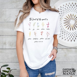 Op de afbeelding: Wit T-shirt met een bloemenontwerp en de tekst "The fruit of the spirit is" gevolgd door een lijst met deugden: liefde, vreugde, vrede, geduld, vriendelijkheid, goedheid, trouw, zachtmoedigheid en zelfbeheersing. De tekst "Galatians 5:22-23" is ook inbegrepen.