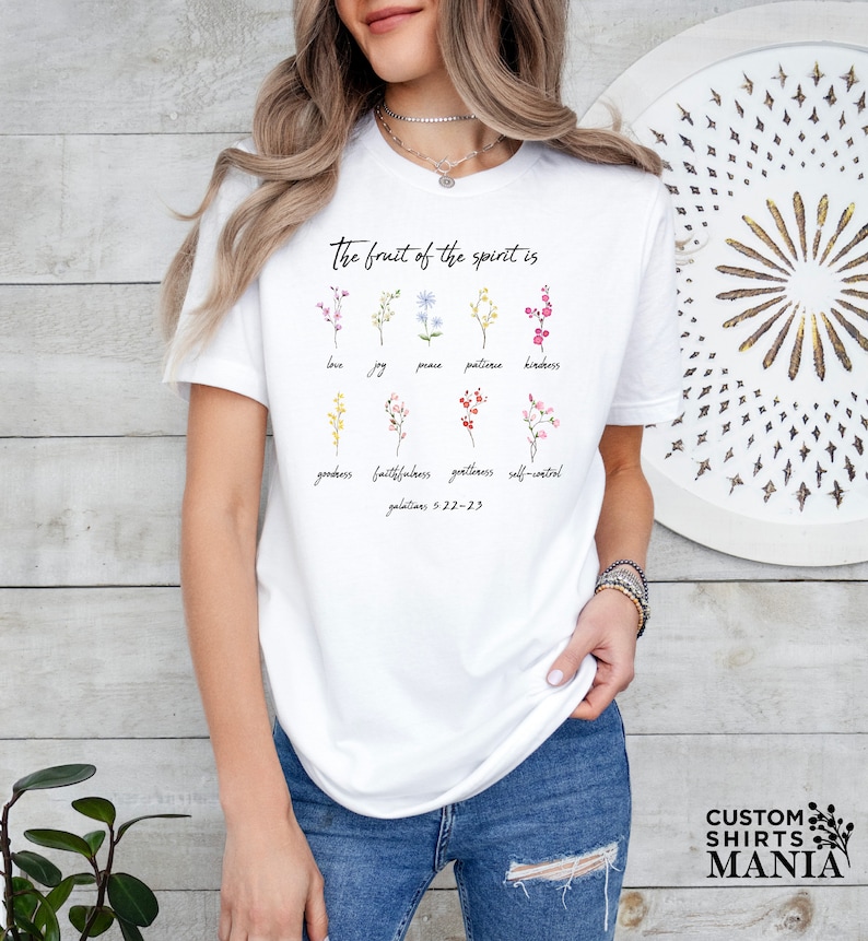 Op de afbeelding: Wit T-shirt met een bloemenontwerp en de tekst "The fruit of the spirit is" gevolgd door een lijst met deugden: liefde, vreugde, vrede, geduld, vriendelijkheid, goedheid, trouw, zachtmoedigheid en zelfbeheersing. De tekst "Galatians 5:22-23" is ook inbegrepen.