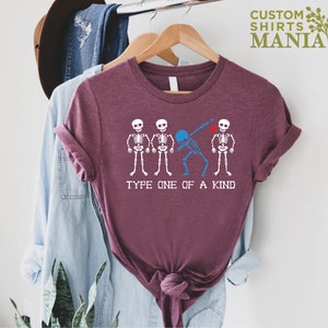 以下が含まれることがあります： 4 つの骸骨のグラフィックが描かれたマルーンのTシャツ。真ん中の骸骨は青色で、ダブをしています。「Type one of a kind」というテキストが骸骨の下にあります。
