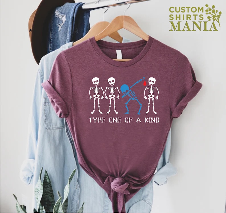 以下が含まれることがあります： 4 つの骸骨のグラフィックが描かれたマルーンのTシャツ。真ん中の骸骨は青色で、ダブをしています。「Type one of a kind」というテキストが骸骨の下にあります。