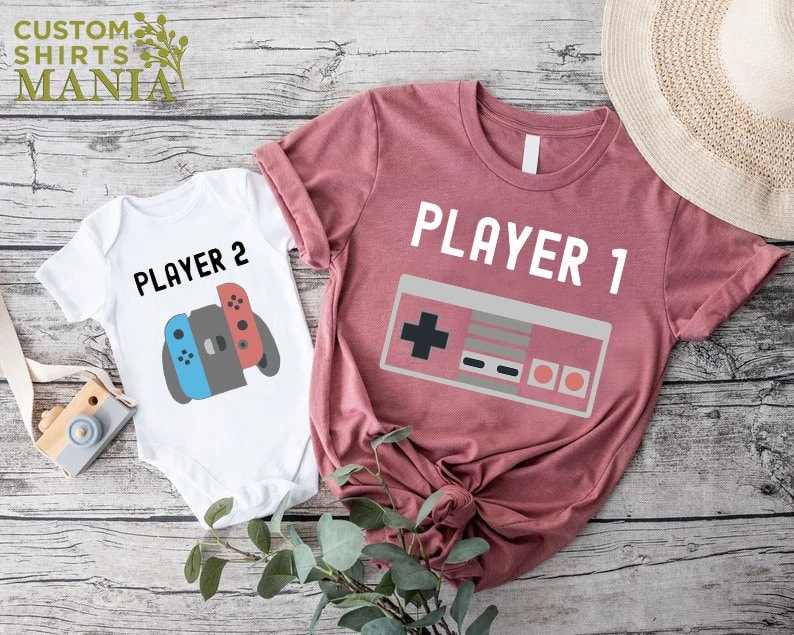 May include: Two white t-shirts with a video game theme. One shirt says "Player 1" with a retro video game controller graphic. The other shirt says "Player 2" with a Nintendo Switch controller graphic.