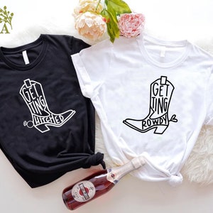 May include: Two white t-shirts with black text and a graphic of a cowboy boot. The text on the black shirt says "Getting Hitched" and the text on the white shirt says "Getting Rowdy".