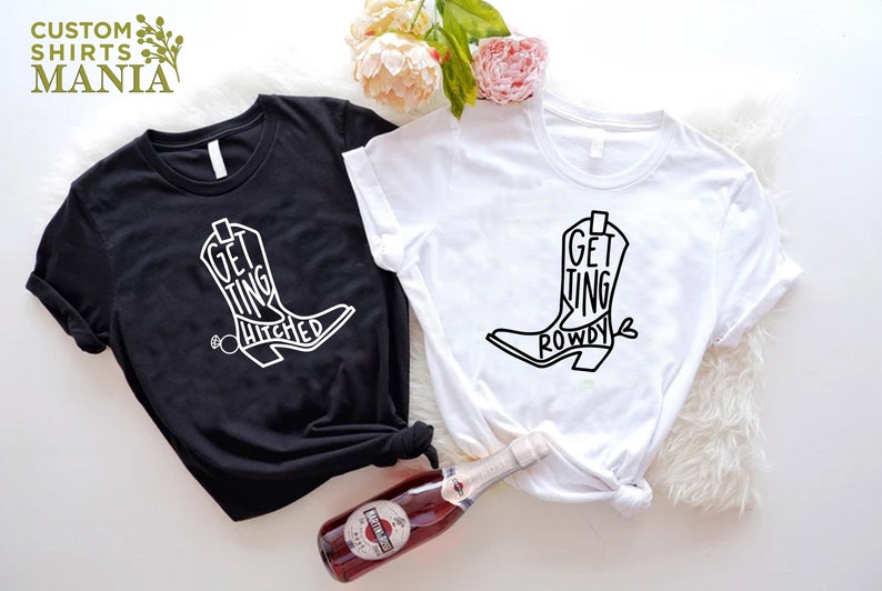 May include: Two white t-shirts with black text and a graphic of a cowboy boot. The text on the black shirt says "Getting Hitched" and the text on the white shirt says "Getting Rowdy".