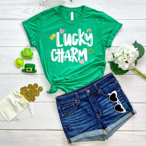 May include: A green t-shirt with the text "Lucky Charm" in white lettering. The shirt has a rainbow, shamrock, horseshoe, and heart graphic. The shirt is lying on a white wooden surface with a pair of blue denim shorts, a bag of gold coins, and a green shamrock.