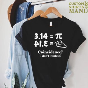 K&ouml;nnte beinhalten: Schwarzes T-Shirt mit wei&szlig;em Text, der "3.14 = &pi;, &pi;.1.&epsilon; =" lautet, mit einer Zeichnung eines Tortenst&uuml;cks. Der Text darunter lautet "Zufall? (Ich glaube nicht)"