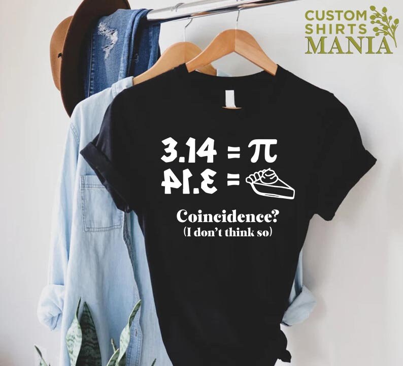 K&ouml;nnte beinhalten: Schwarzes T-Shirt mit wei&szlig;em Text, der "3.14 = &pi;, &pi;.1.&epsilon; =" lautet, mit einer Zeichnung eines Tortenst&uuml;cks. Der Text darunter lautet "Zufall? (Ich glaube nicht)"