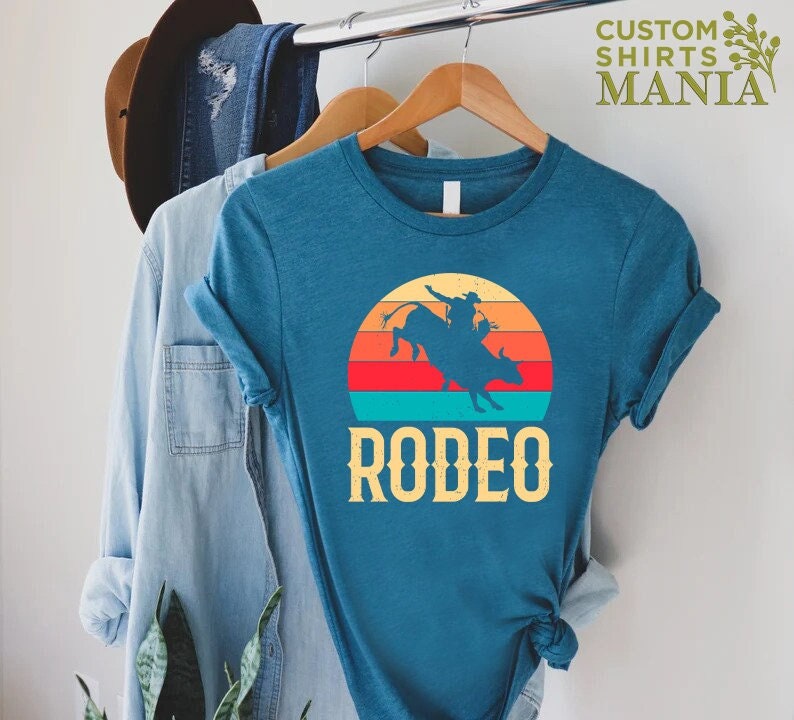 May include: A teal blue t-shirt with a vintage-style sunset graphic of a cowboy riding a bull. The text "RODEO" is printed below the graphic.