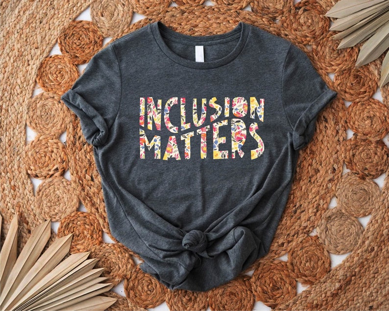 Puede incluir: Una camiseta gris oscuro con el texto "Inclusion Matters" impreso en letras blancas con un patr&oacute;n floral. La camiseta est&aacute; colocada sobre una estera de paja tejida.