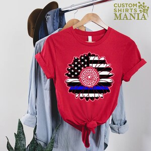 May include: Red t-shirt featuring a black and white sunflower design. The sunflower is made up of American flag stripes with a blue line running through the centre.