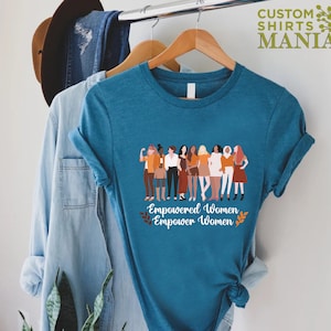 May include: Teal blue t-shirt featuring a graphic of a group of diverse women standing together. The text "Empowered Women Empower Women" is printed below the graphic.