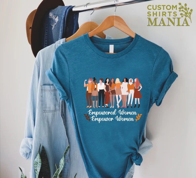 May include: Teal blue t-shirt featuring a graphic of a group of diverse women standing together. The text "Empowered Women Empower Women" is printed below the graphic.