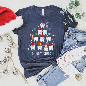以下が含まれることがあります： 歯でできたクリスマスツリーのデザインが施されたネイビーブルーのTシャツ。ツリーはサンタの帽子、キャンディーケーン、プレゼントで飾られています。ツリーの下には「Oh Dentistree」という文字が印刷されています。