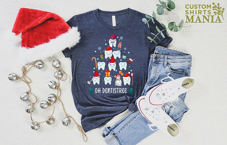 以下が含まれることがあります： 歯でできたクリスマスツリーのデザインが施されたネイビーブルーのTシャツ。ツリーはサンタの帽子、キャンディーケーン、プレゼントで飾られています。ツリーの下には「Oh Dentistree」という文字が印刷されています。