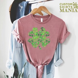 May include: A pink t-shirt with a green four-leaf clover design made of smaller clovers. The design is centered on the shirt. The shirt is hanging on a wooden hanger.