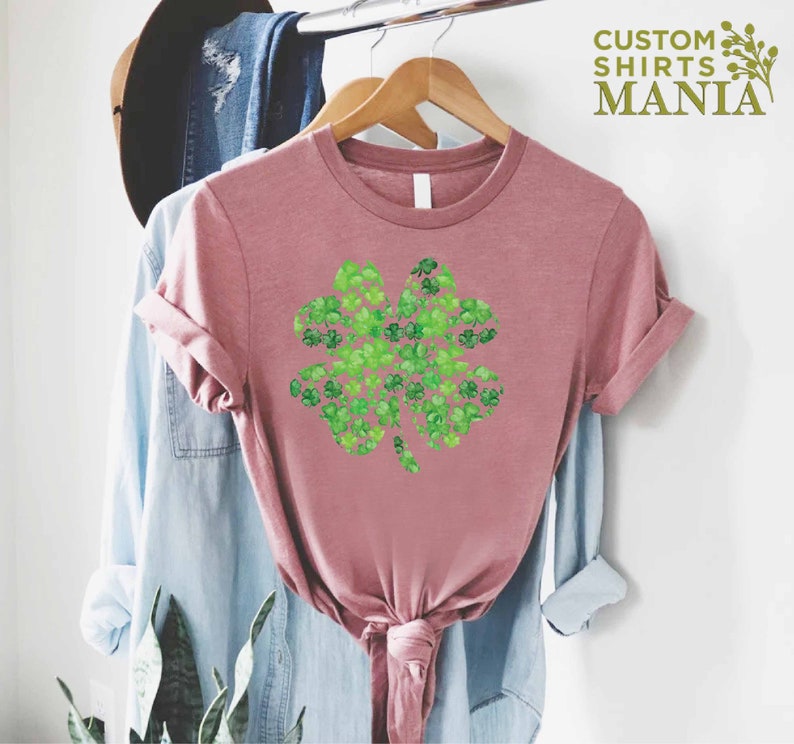 May include: A pink t-shirt with a green four-leaf clover design made of smaller clovers. The design is centered on the shirt. The shirt is hanging on a wooden hanger.