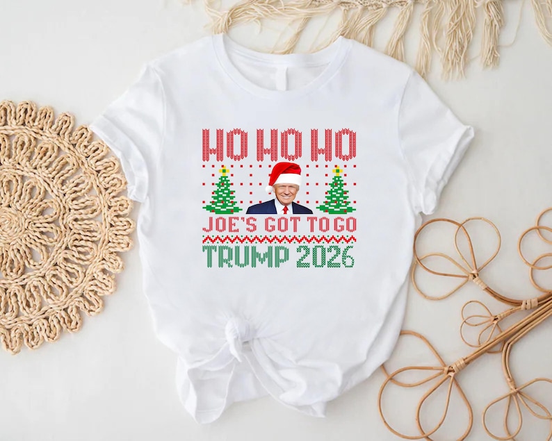Op de afbeelding: Een wit T-shirt met korte mouwen en een grafische print in rood, groen en blauw. Het ontwerp bevat de tekst HO HO HO, JOE'S GOT TO GO en TRUMP 2026, samen met twee groene kerstbomen en een portret van Donald Trump met een kerstmuts op.