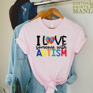 K&ouml;nnte beinhalten: Rosa T-Shirt mit dem Text "ICH LIEBE jemanden mit AUTISMUS" in Regenbogenfarben. Ein Puzzle-Herz befindet sich &uuml;ber dem Wort "LOVE".