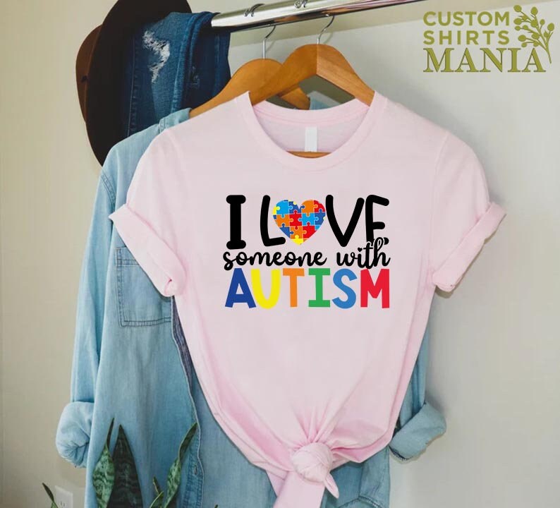K&ouml;nnte beinhalten: Rosa T-Shirt mit dem Text "ICH LIEBE jemanden mit AUTISMUS" in Regenbogenfarben. Ein Puzzle-Herz befindet sich &uuml;ber dem Wort "LOVE".