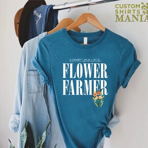May include: A teal blue t-shirt with white text that reads "Support Your Local Flower Farmer". The text is centered on the shirt and is surrounded by a small bouquet of flowers.