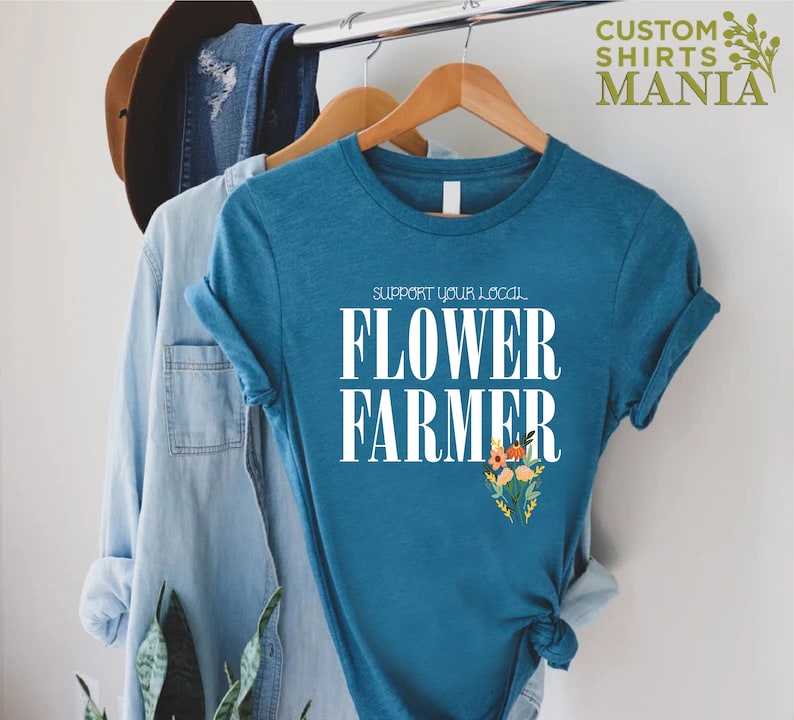 May include: A teal blue t-shirt with white text that reads "Support Your Local Flower Farmer". The text is centered on the shirt and is surrounded by a small bouquet of flowers.