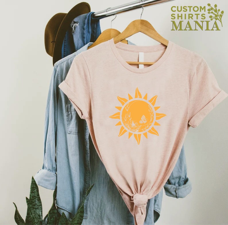 Puede incluir: Una camiseta rosa p&aacute;lido con un gr&aacute;fico de sol amarillo que presenta una mariposa y detalles florales. El texto "CUSTOM SHIRTS MANIA" es visible en la esquina superior derecha de la imagen.