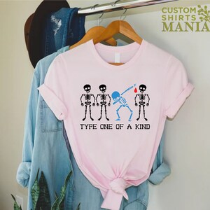以下が含まれることがあります： ピンクのTシャツに、4つの骸骨のグラフィックがプリントされています。1つの骸骨はダブをしていて、体は青色です。骸骨の下には「Type One of a Kind」という文字がプリントされています。