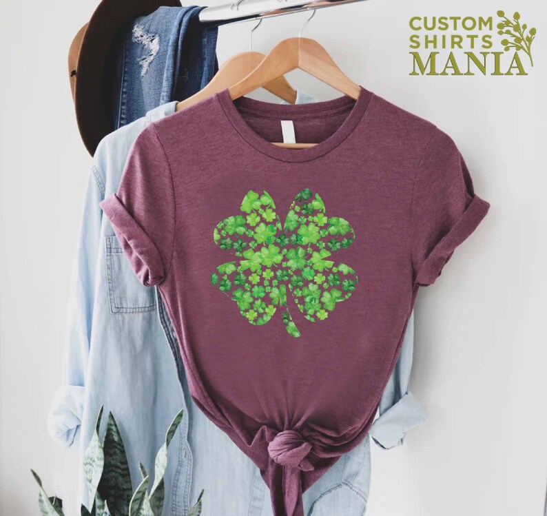 May include: A maroon t-shirt with a green four-leaf clover design made of smaller clovers. The design is centered on the shirt. The shirt is hanging on a hanger.