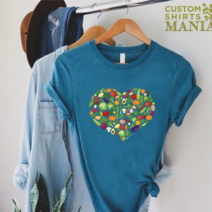 May include: Teal t-shirt featuring a heart-shaped design composed of various colorful vegetables. The shirt includes the text "CUSTOM SHIRTS MANIA" in green. The vegetables include tomatoes, peppers, and avocados.