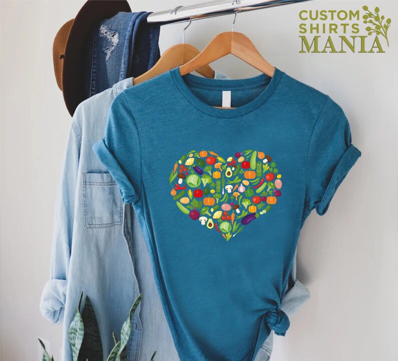May include: Teal t-shirt featuring a heart-shaped design composed of various colorful vegetables. The shirt includes the text "CUSTOM SHIRTS MANIA" in green. The vegetables include tomatoes, peppers, and avocados.