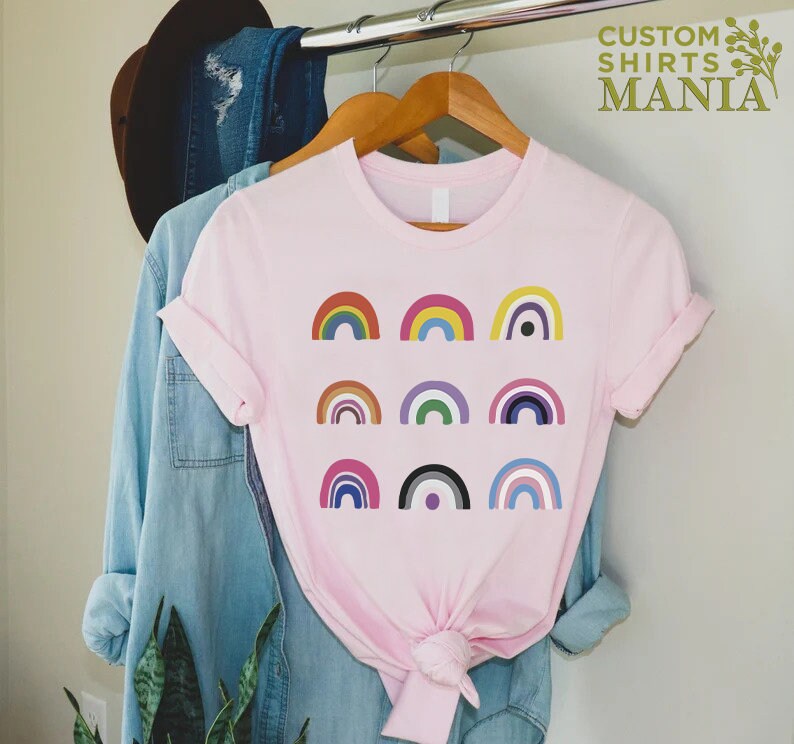 K&ouml;nnte beinhalten: Ein pinkfarbenes T-Shirt mit einem Design aus neun verschiedenen bunten Regenb&ouml;gen. Die Regenb&ouml;gen sind in drei Reihen zu je drei angeordnet. Der Text "CUSTOM SHIRTS MANIA" ist in der oberen rechten Ecke des Bildes sichtbar.