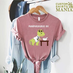May include: A pink t-shirt featuring the text "PHARMASAURUS RX" and a cartoon dinosaur wearing glasses and a lab coat holding a mortar and pestle.