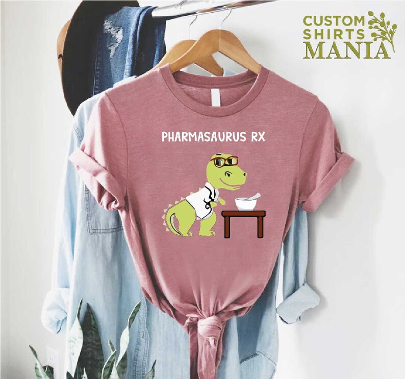 May include: A pink t-shirt featuring the text "PHARMASAURUS RX" and a cartoon dinosaur wearing glasses and a lab coat holding a mortar and pestle.