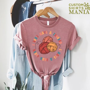 May include: A pink t-shirt with the text "I don't have a problem I have a yarn stash" in a colorful font. The text is surrounded by a circle with three balls of yarn in the center.
