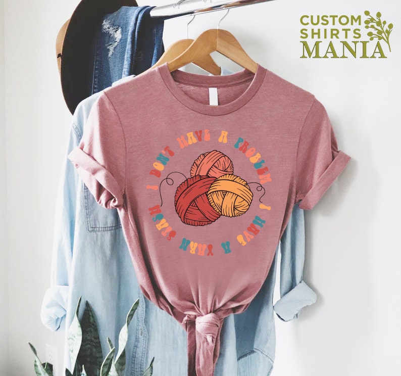 May include: A pink t-shirt with the text "I don't have a problem I have a yarn stash" in a colorful font. The text is surrounded by a circle with three balls of yarn in the center.
