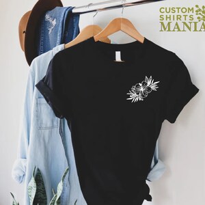 May include: Black t-shirt with a white floral design on the left chest. The shirt is hanging on a wooden hanger.