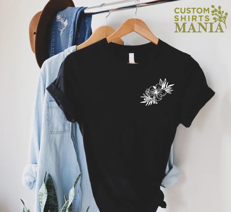 May include: Black t-shirt with a white floral design on the left chest. The shirt is hanging on a wooden hanger.