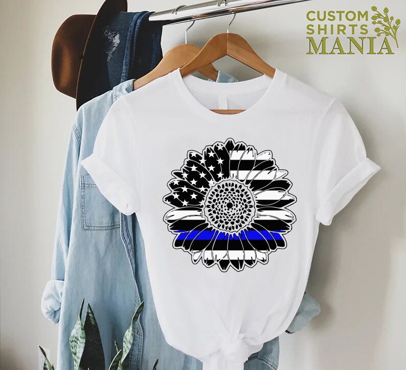 May include: White t-shirt featuring a black and white sunflower design. The sunflower has a blue line running through the centre, representing the thin blue line.