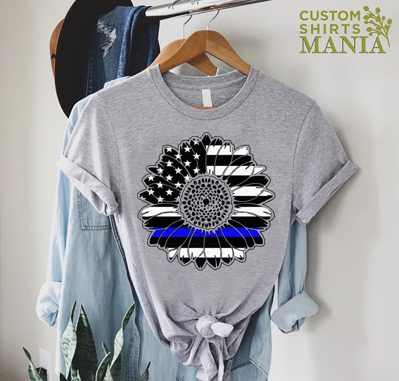 May include: A grey t-shirt featuring a black and white sunflower design. The sunflower is made up of the American flag with a blue line running through the centre.