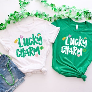 May include: Two t-shirts with the text "Lucky Charm" in green lettering with colourful accents. The shirts are white and green.