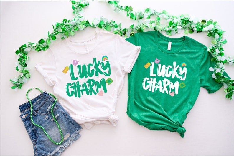 May include: Two t-shirts with the text "Lucky Charm" in green lettering with colourful accents. The shirts are white and green.