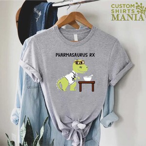 May include: A grey t-shirt featuring a cartoon dinosaur wearing glasses and a lab coat. The dinosaur is holding a mortar and pestle. The text "PHARMASAURUS RX" is printed on the shirt.